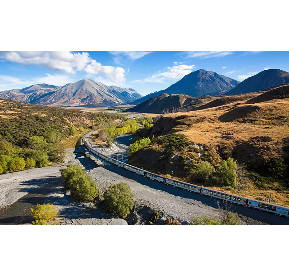 Kiwirail Great Journeys of New Zealand Tranzalpine train travels along the banks of the river with mountains behind