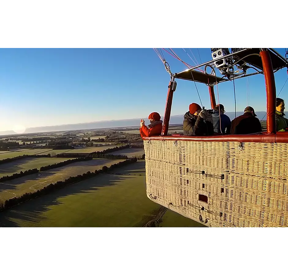 Tourists seen in the basket of the balloon while in flight, taking photos of the view. The day is clear with blue skies. The landscape is lush green Canterbury plains