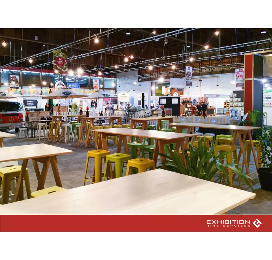 Exhibition Hire Services Trade Show, wooden bar leaners with colored bar stools. In the background trade stands are set up